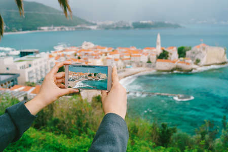 Girls hands hold a smartphone with a photo of buildings on the background of the city of Budvaの写真素材