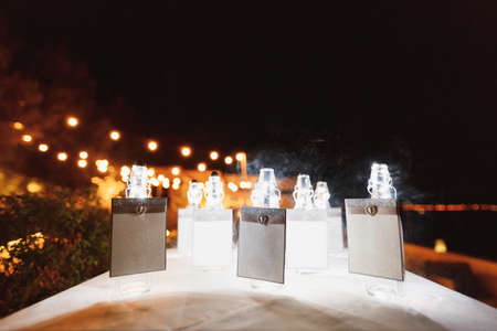 Sealed bottles with blank signs stand on a table covered with a white tablecloth against a backdrop of glowing light bulbs at nightの写真素材