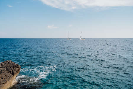 Two white sailboats are sailing on the open sea on a sunny bright day. View from the rocky shoreの写真素材