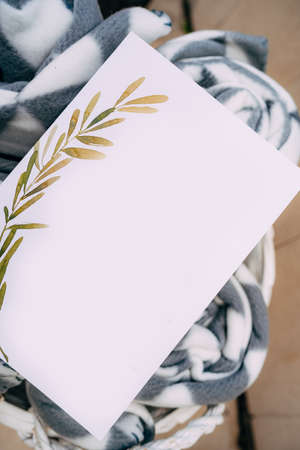 Blank invitation with an ornament lies on a rolled up plaid blanket in a white wicker basket