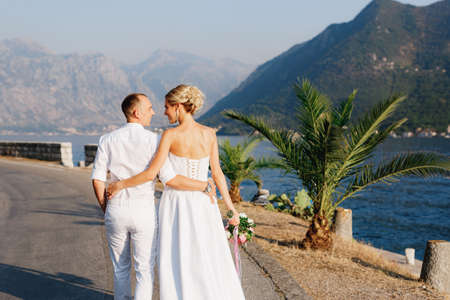 The bride and groom walk along the pier near the old town of Perast, hugging each other, back view