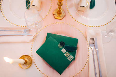 Blank green envelopes with stamps lie on a transparent plate on a set table. A lighted candle is...