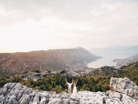 The bride and groom stand on Mount Lovcen with their hands up and look at the Bay of Kotorの写真素材