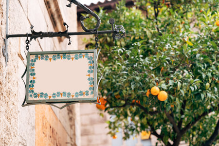 Beautiful blank board of a fancy shape hangs in a forged frame on the facade of a building against a background of a tangerine treeの写真素材