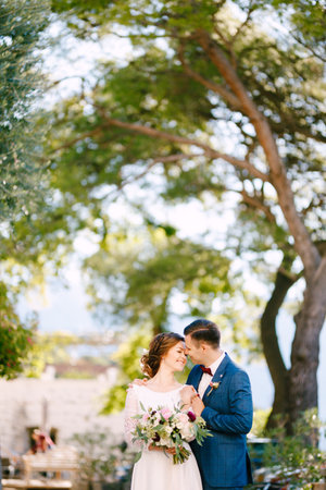Groom gently hugs the bride by the shoulders against the backdrop of trees on a warm sunny day. Bride holds a beautiful bouquet of flowers in her handsの写真素材