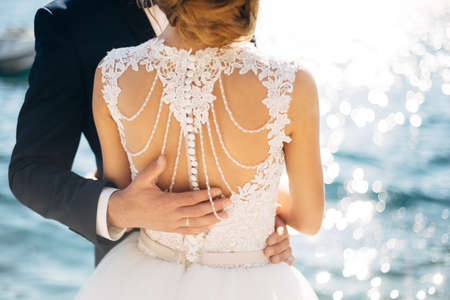 The bride and groom are standing and hugging on the pier near the sea, hand of the groom on brides back, close-upの写真素材