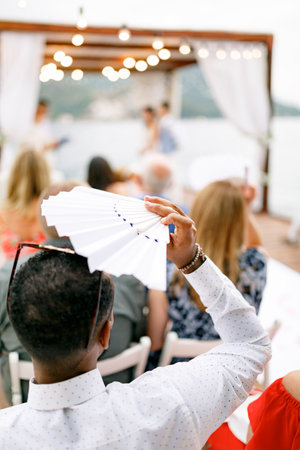 The guest at the wedding ceremony sits, covers from the sun and fancies himself with a white fan, back viewの写真素材