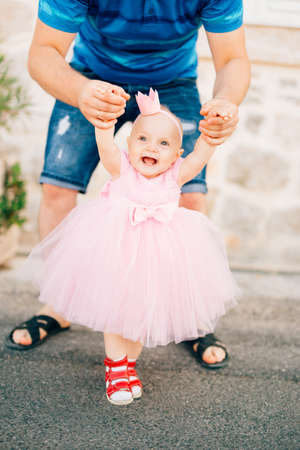 A charming little girl in a pink dress, sneakers and with a crown on her head holding dad by the hands and laughingの写真素材