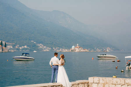 The bride and groom are embracing on the pier near the old town of Perast, boats are floating nearby