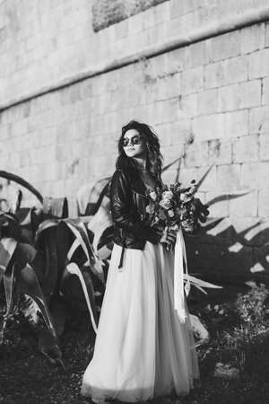 Black and white photo of bride in sunglasses with a bouquet of flowers against the background of the castle wall and agaveの写真素材