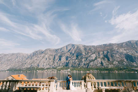 Newlyweds stand embracing near the ancient fence on the shore against the background of mountains and the seaの写真素材