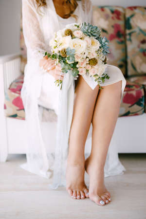 Barefoot bride in a lace robe sits on a sofa in a hotel room and holds a wedding bouquet in her hands, close-upの写真素材