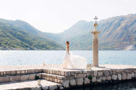 A bride walks along a stone pier next to an old column in the Bay of Kotor, her skirt flutters in the windの写真素材