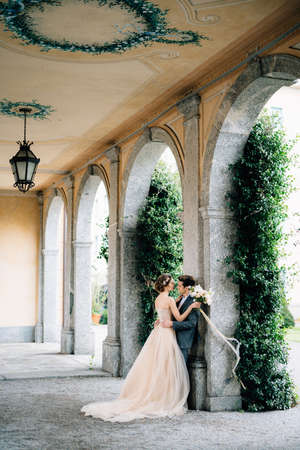 Bride with a bouquet of flowers embraces groom leaning against an arch entwined with green ivy. Lake Comoの写真素材