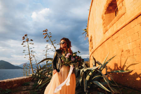 Bride in a pastel dress and sunglasses holds a bouquet of flowers in her hands stands against the background of blooming agave bushes and the walls of the castleの写真素材