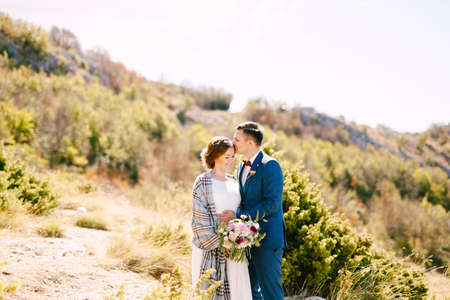Groom kissing bride in a checkered shawl with a bouquet of flowers in her hands on the background of rocks and green plantsの写真素材