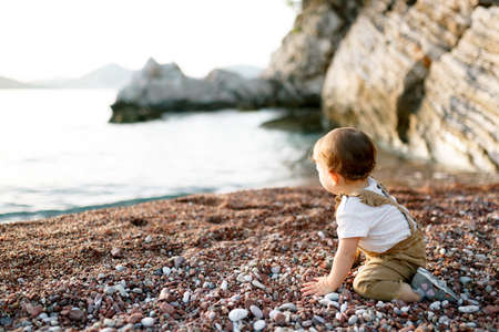 Little boy sits on a pebble beach by the sea and looks into the distanceの写真素材