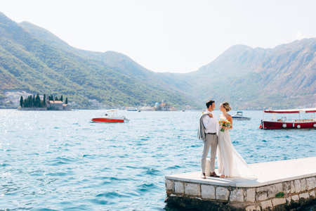 The bride and groom standing side by side on the pier in the Bay of Kotor, islands of Perast are behind themの写真素材