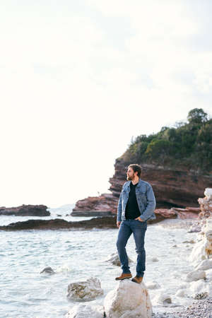 Man in jeans and a denim jacket stands looking at the sea on a large stone on a pebble beach against...
