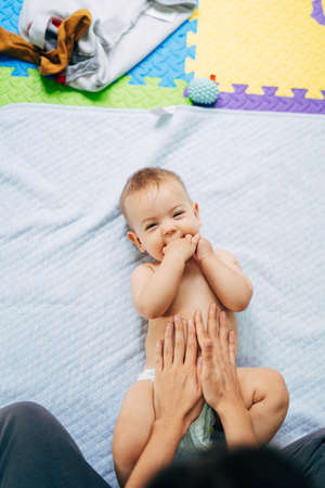 Smiling baby in a diaper with his hands in his mouth lies with his back on a blue blanket on a...