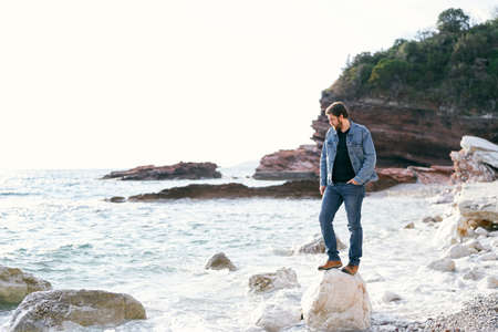 Man in jeans and a denim jacket stands looking under his feet on a large stone on a pebble beach against a background of green rocky mountainsの写真素材