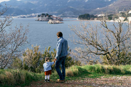 Dad holds the hand of a little girl, standing on the coast against the background of the sea,...