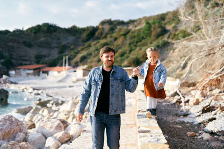 Smiling dad leads a little girl by the hand along the curb on a pebble beach against a background of...
