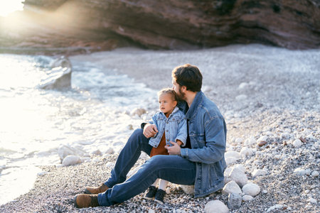 Dad sits on a stone, hugging and kissing the top of the head of a little girl on a pebble beach by the sea against the backdrop of rocksの写真素材