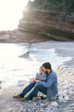 Dad with his little daughter sits on a pebble beach near the sea against the backdrop of rocky mountainsの写真素材
