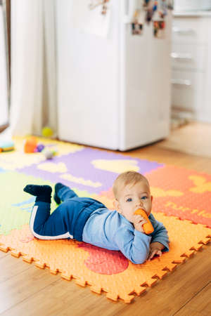 Little baby boy in a blue suit with a carrot in his hands lies on his tummy on a colored rug on the...