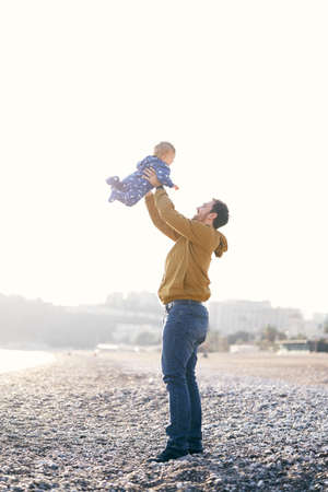 Smiling dad in a brown jacket and jeans throws a small child in a blue overalls high on his arms while standing on a pebble beachの写真素材
