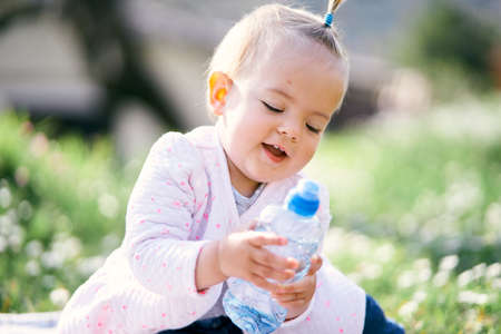 Smiling little girl with a ponytail on her head sits on a green lawn and holds a bottle in her handsの写真素材