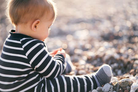 Kid in a striped jumpsuit sits on a pebble beach, holding his hands in front of him. Back viewの写真素材