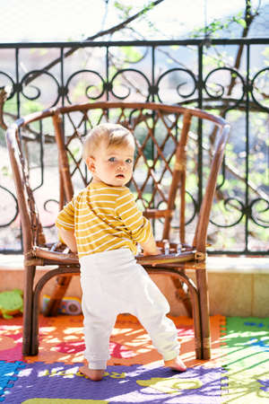 Kid stands on the balcony near the wicker chair, turning his head back