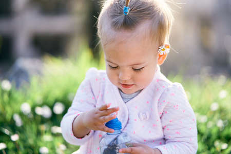 Little baby with a chamomile behind her ear sits on a green lawn and opens a plastic bottle....