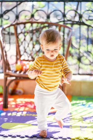 Small child walks on a colored rug against the background of a wicker chair on the balconyの写真素材