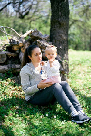 Mom holds a little girl in her arms, sitting in the park near felled stumpsの写真素材