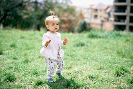 Little girl stands on a green meadow among the trees on the background of buildingsの写真素材