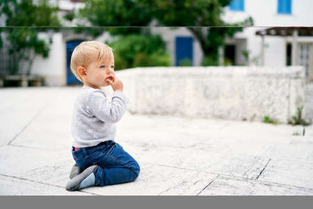 Pensive kid sits on his knees on a tile on the background of a buildingの写真素材