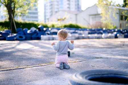 Child is kneeling against the background of car tires in the parking lot. Back viewの写真素材