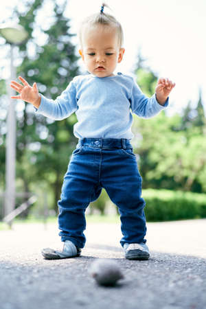 Little girl with a ponytail stands on the road in the park and looks at a pebbleの写真素材