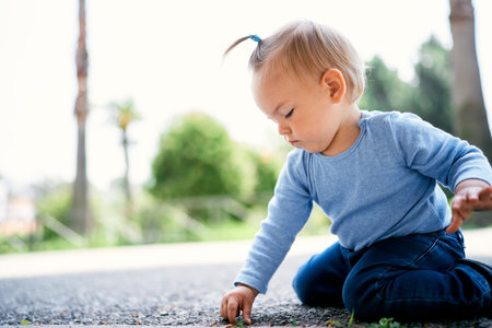 Little girl sits on her lap and picks the asphalt in a green park. Side viewの写真素材