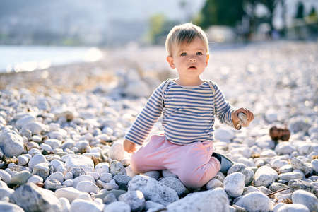 Baby sits on his knees on a pebble beach and holds a pebble in his hand