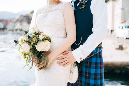 A bride in a wedding dress with bridal bouquet and a groom in a Scottish national dress stand hugging on the pierの写真素材