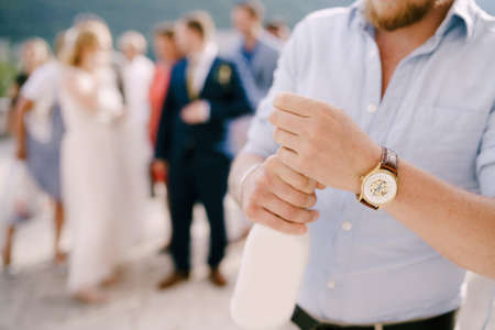 A man at a wedding ceremony opens a bottle of champagne, behind him guests and newlyweds, close-upの写真素材