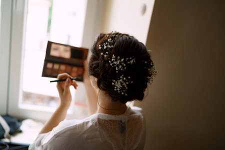 Bride applies makeup while standing by the window in a hotel room before the wedding ceremony, back viewの写真素材