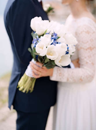 The bride and groom stand hugging and hold a wedding bouquet with white and blue flowers and eucalyptus branches close-upの写真素材