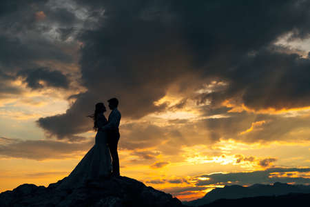 The bride and groom are embracing on the rocks in the mountains at sunset, silhouettesの写真素材