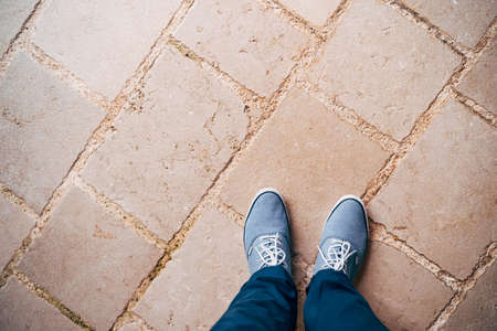 Legs of a man in blue pants and blue sneakers standing on the stone road, top view