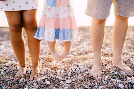 Legs of mom, dad and little daughter in a dress standing on a pebble beachの写真素材
